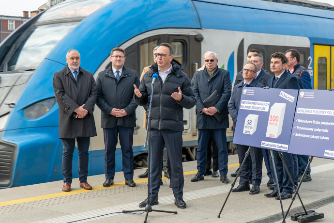 Minister Infrastruktury podczas briefingu prasowego. W tle pociąg.
